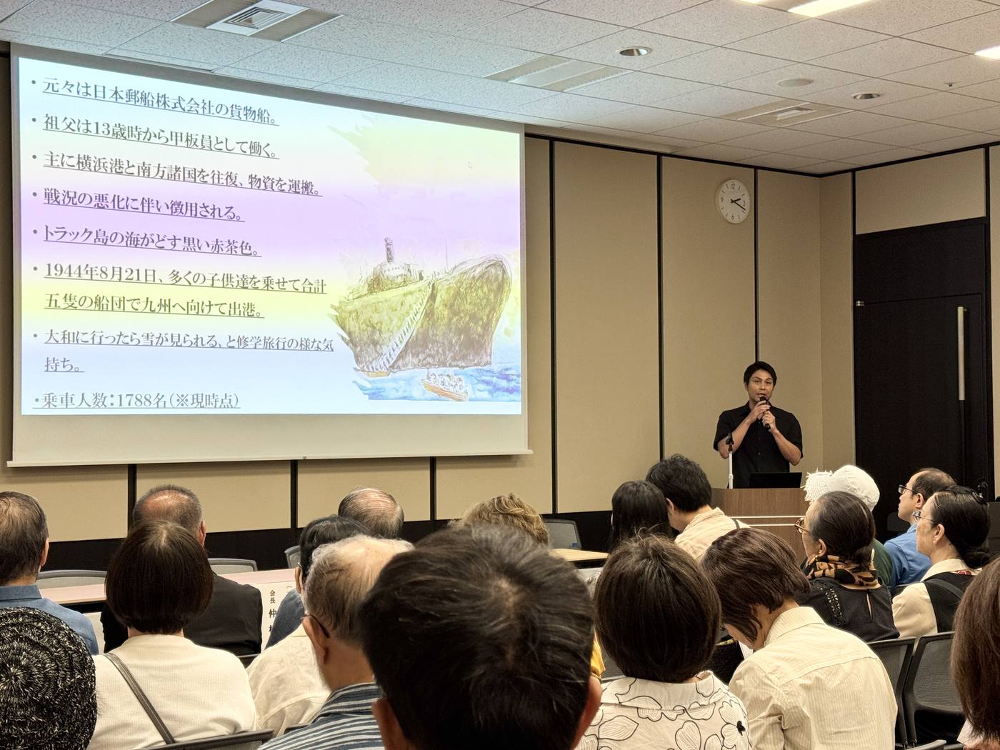 対馬丸となった貨物船の来歴をまとめたスライドを背に、マイクを持って講演する監督・寿大聡