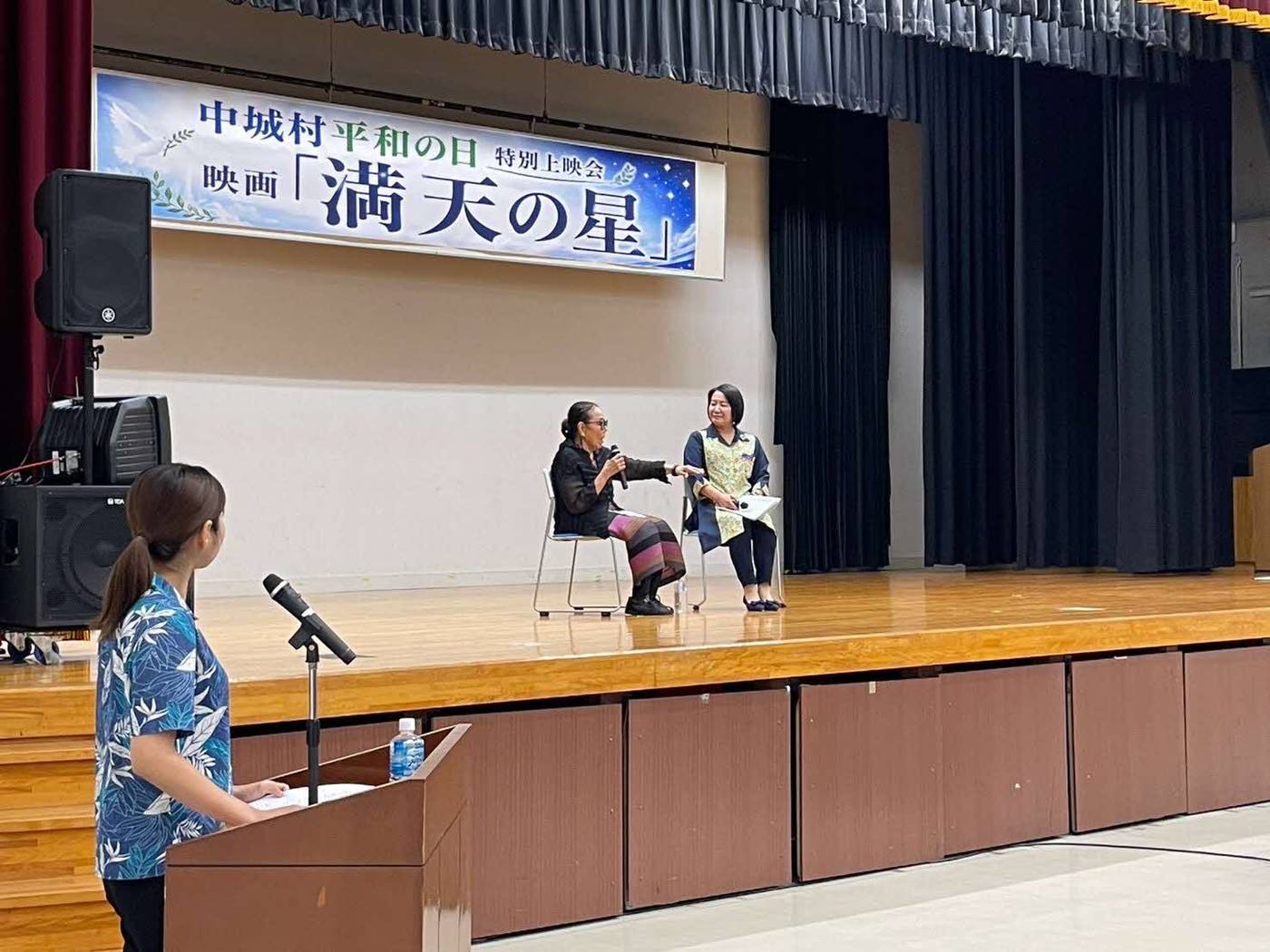 「中城村平和の日 特別上映会 映画『満天の星』」の横断幕を掲げたステージで、マイクを持って話すトークセッションの登壇者